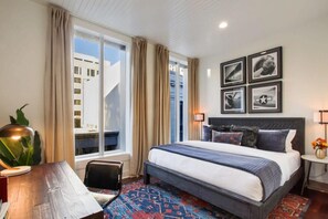 Een hotelkamer met een groot bed, een bureau met lamp, een stoel, een raam met gordijnen en ingelijste foto's aan de muur.