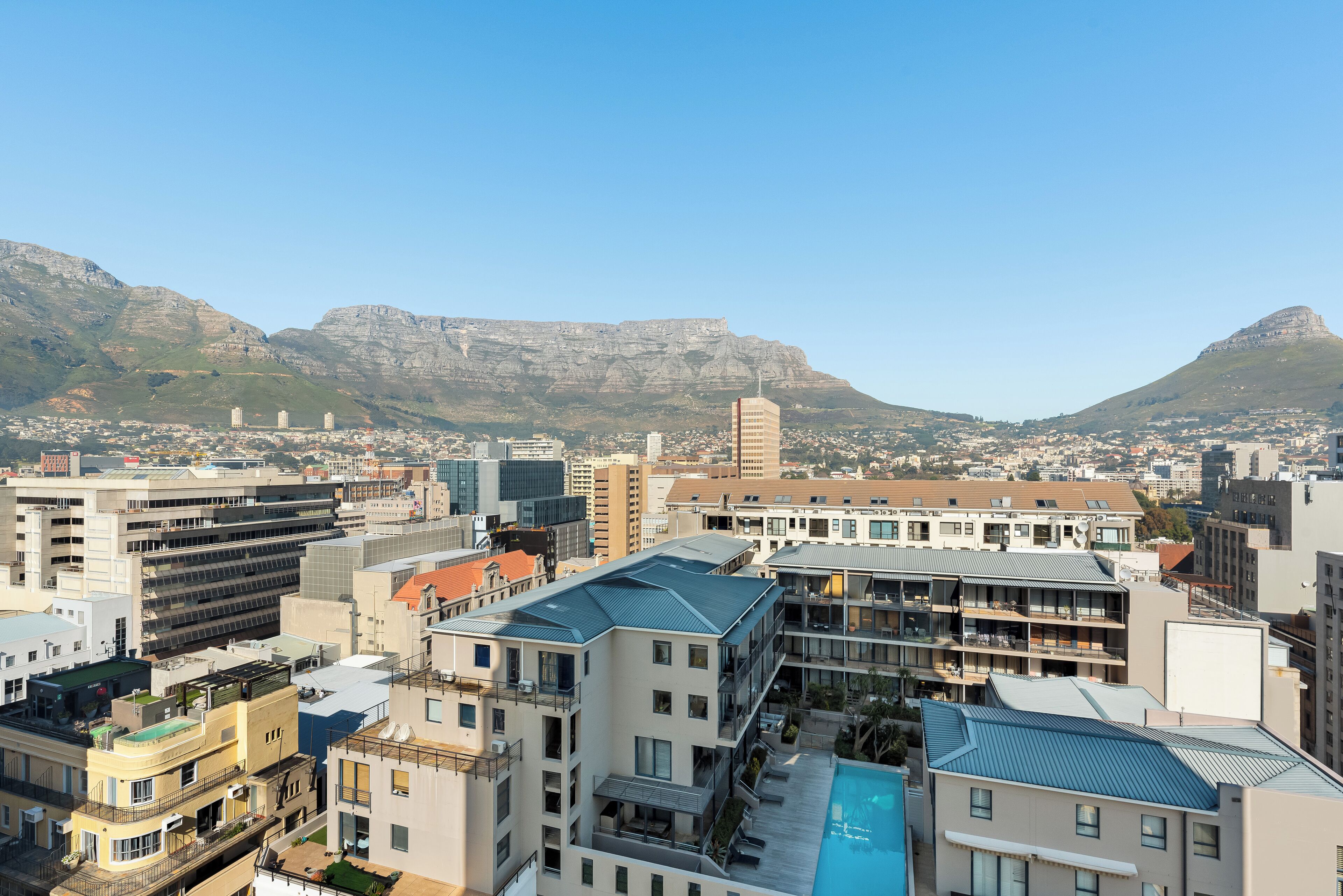 Un paisaje urbano con edificios modernos, un hotel destacado con piscina en la azotea y una cadena montañosa al fondo.