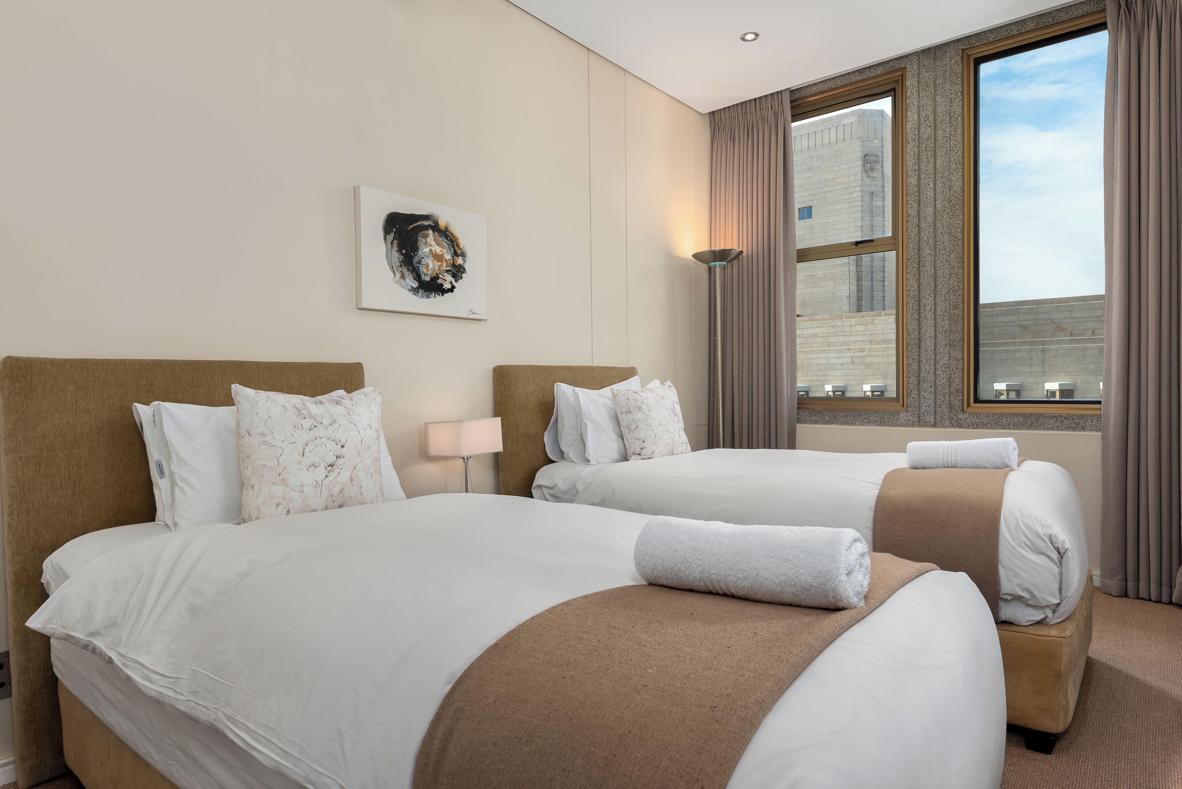 A hotel room with two beds, a large window, a painting on the wall, and a lamp.