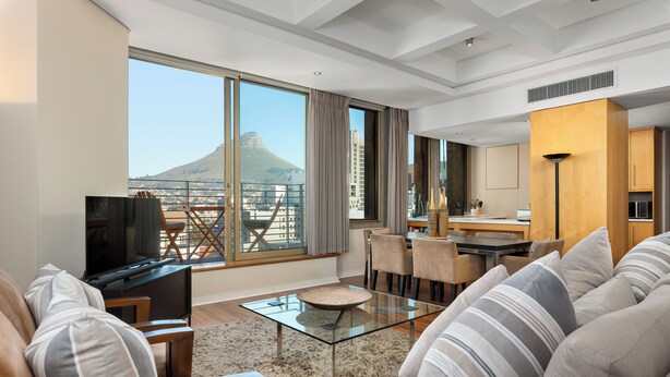 A modern living room with a glass coffee table, sofas, and a TV. A balcony with a city view and a mountain in the distance is visible. - Cartwrights Corner 1403 (Cape Town)