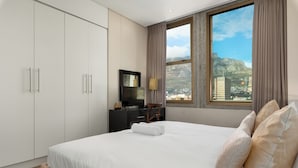 A hotel room with a large bed, a TV, a desk, and a view of a cityscape through a window. - Cartwrights Corner 1403 (Cape Town)