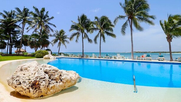 Una piscina azul cristalina rodeada de palmeras y sillas de playa, con una gran roca al frente.