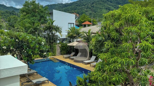 A swimming pool with lounge chairs and a waterfall feature, surrounded by lush greenery and a mountain backdrop. - Hill Myna Holiday Park & Cafe (Choeng Thale)