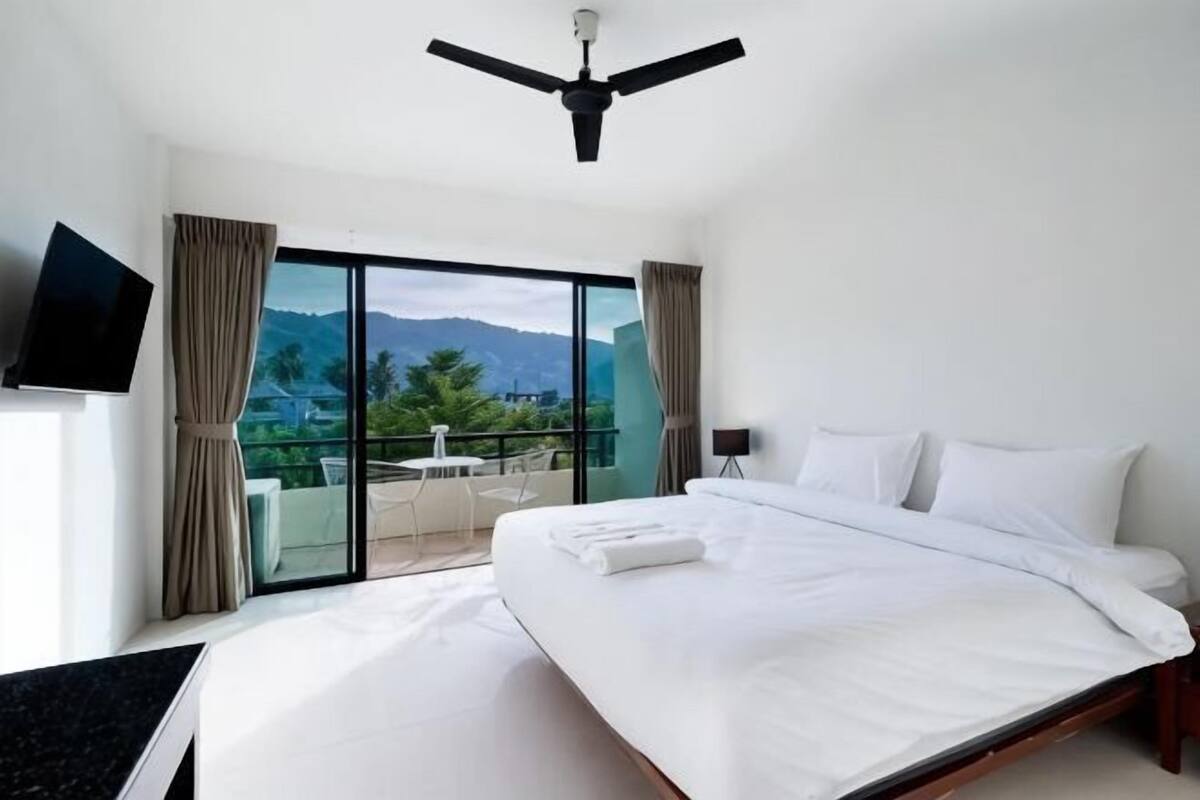 A hotel room with a bed, a ceiling fan, a TV, and a balcony with a view of mountains.