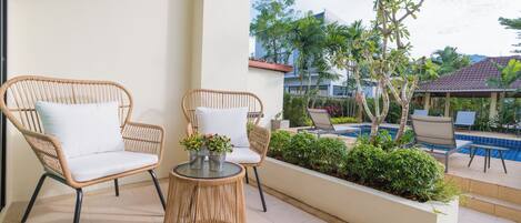 A balcony with wicker furniture, a small table with flowers, and a view of a swimming pool and greenery.