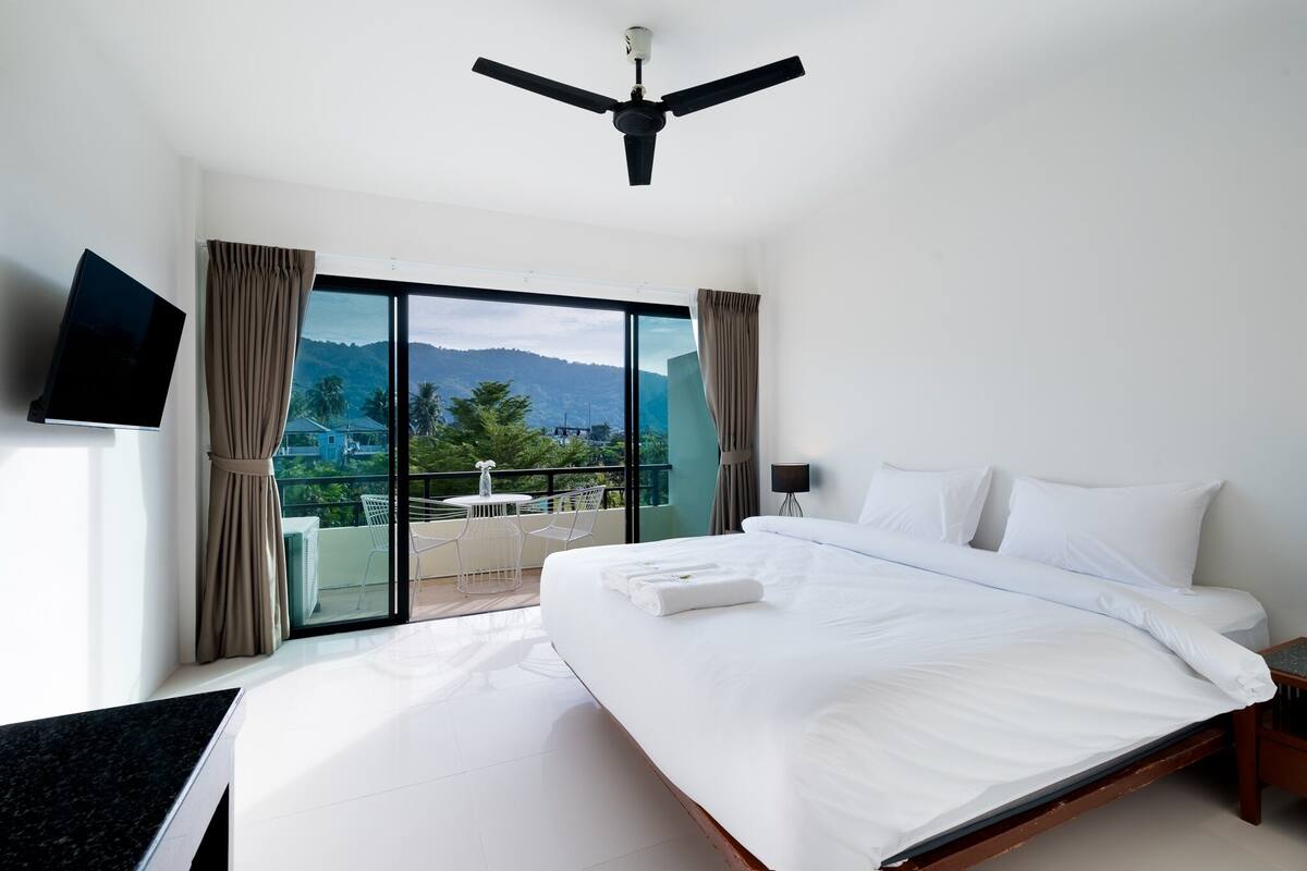 Habitación de hotel con una cama grande, ventilador de techo, televisor y balcón con mesa y sillas.
