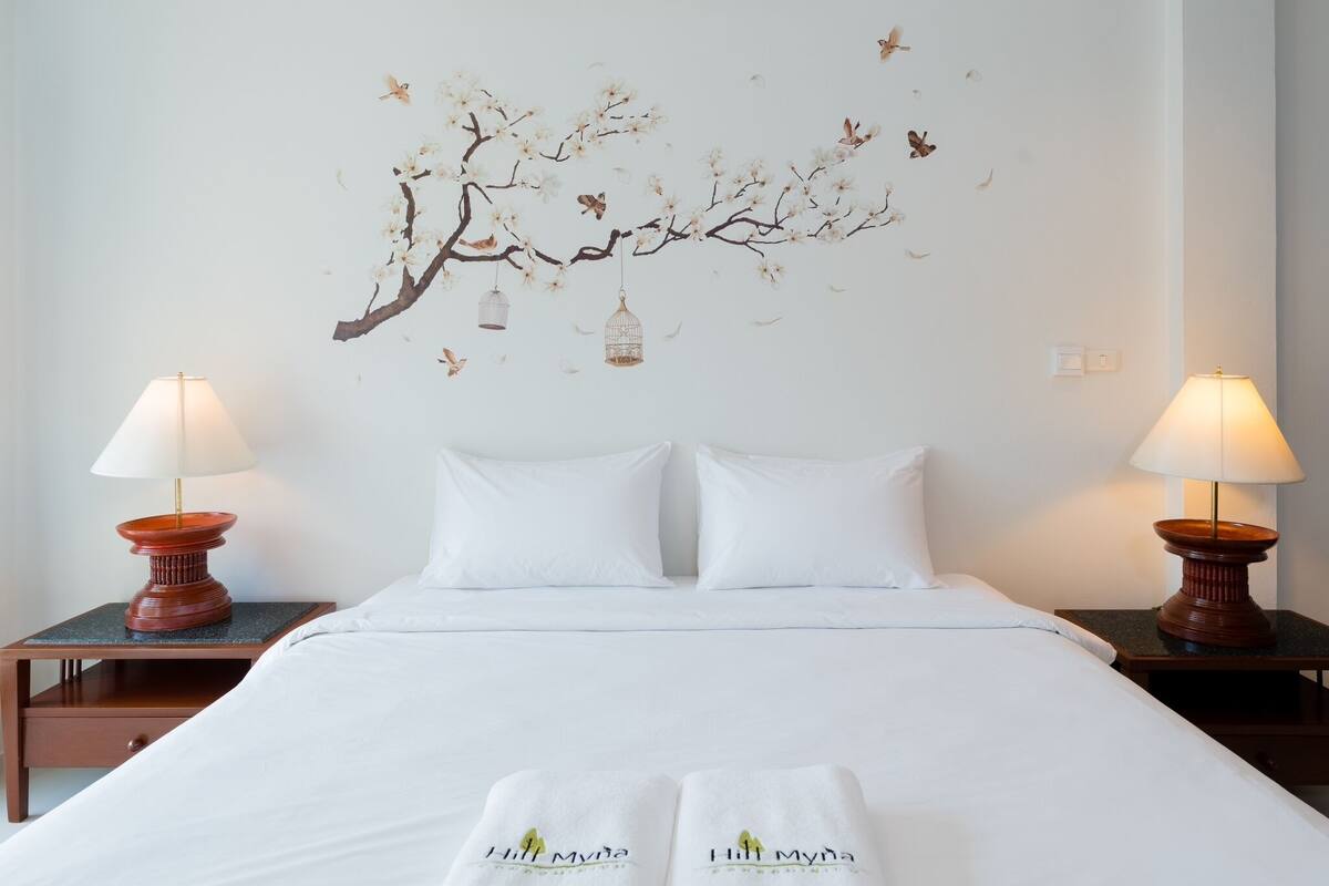 Una cama bien hecha con sábanas blancas y dos almohadas, flanqueada por mesitas de noche de madera con lámparas. Se aprecia en la pared un mural con una rama de árbol, pájaros y mariposas.