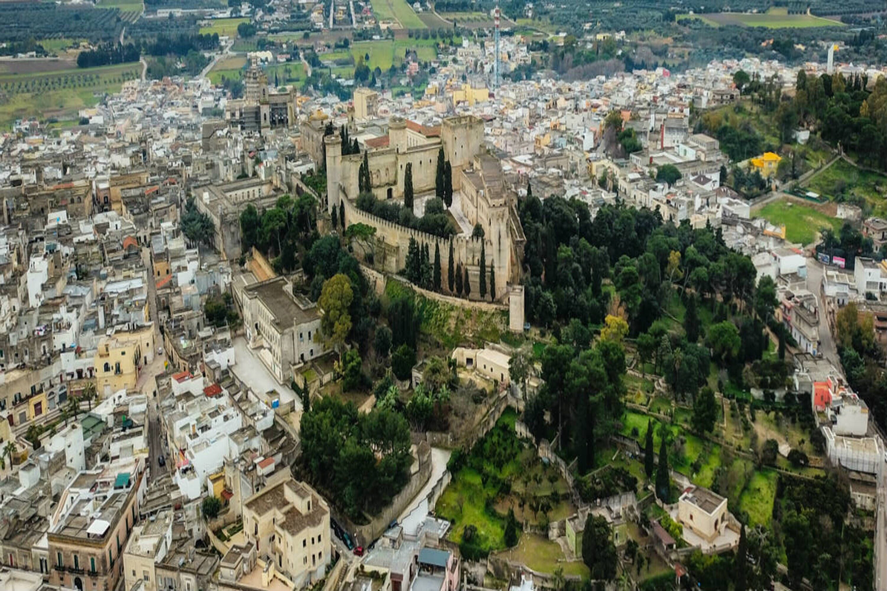 Vista aérea de uma cidade histórica com um castelo imponente, cercada por áreas urbanas densas e vegetação.
