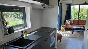 Eine kompakte Küche mit Fenster, Spüle und Herd. Im angrenzenden Wohnbereich befindet sich eine Couch und ein kleiner Hocker.