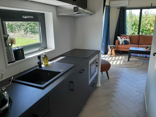 A compact kitchen with a window, a sink, and a stove. There is a sofa and a small stool in the adjacent living area.