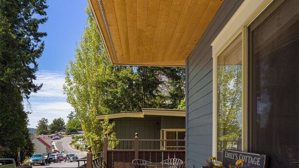 Una terraza de madera con mobiliario exterior, un cartel que dice "EMILY'S COTTAGE" y una vista de una carretera con coches.