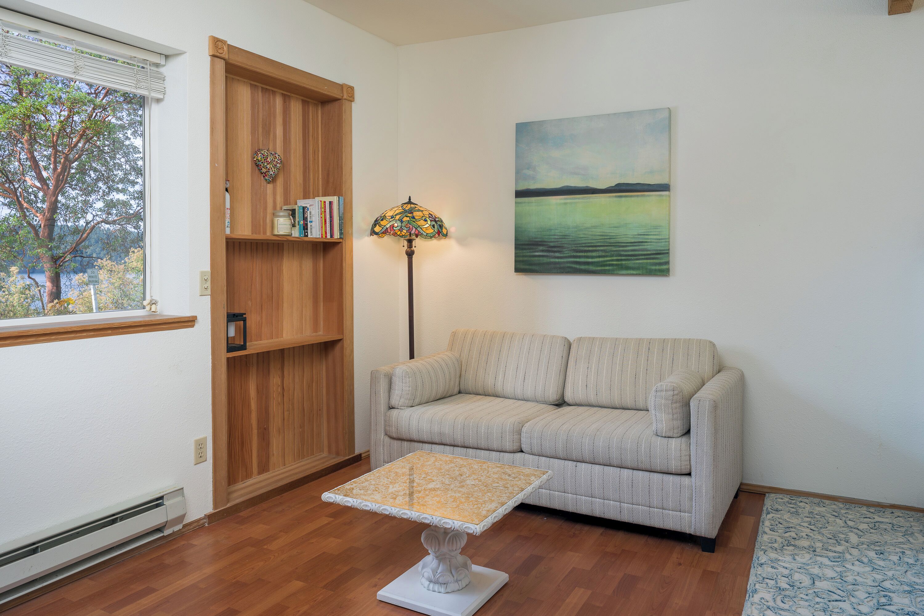 Una sala de estar con un sofá, una estantería de madera, una mesa, una lámpara y un cuadro en la pared.