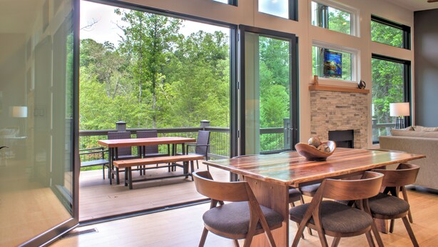 Un comedor moderno con una mesa grande y sillas, una chimenea y ventanas amplias que dan a un bosque.