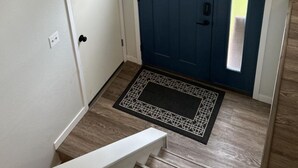Una porta d'entrada amb una porta blava i una finestra, una banca blanca, un felpú i una làmpada penjant a la paret.