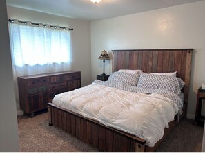 A bedroom with a wooden bed frame, a nightstand with a lamp, a dresser with multiple drawers, and a window with a curtain.