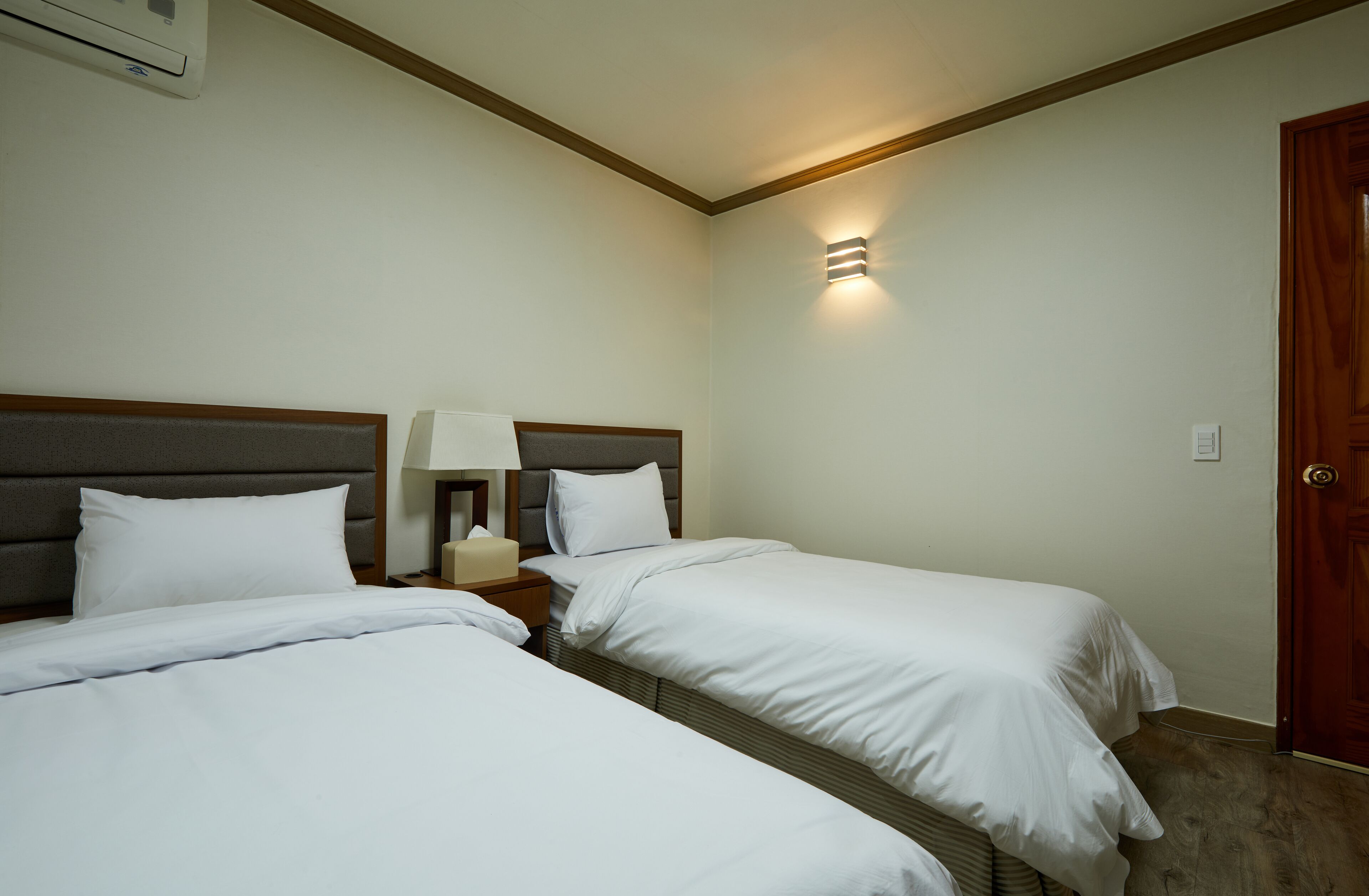 Een hotelkamer met twee bedden, een nachtkastje met een lamp, een wandlamp en een houten deur.