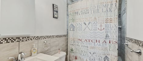 Ein Badezimmer mit weißem Waschbecken, einem beleuchteten Spiegel, Toilette und einer Duschvorhang mit Aufschrift.