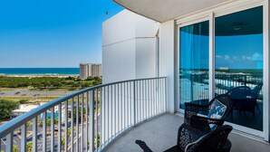 A balcony with a view of the ocean and cityscape, a wicker chair, and a glass door.