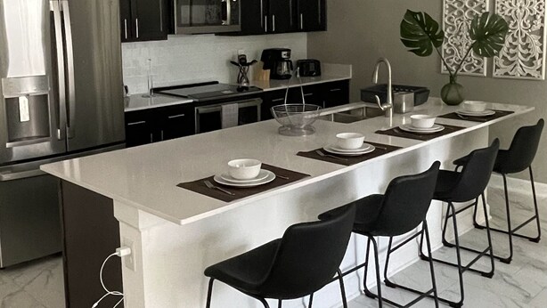 A modern kitchen with dark cabinets, a stainless steel refrigerator, a microwave, and a large island with bar stools. - 1094ssp-the Retreat At Championsgate 4 Bedroom Home by RedAwning (Davenport)