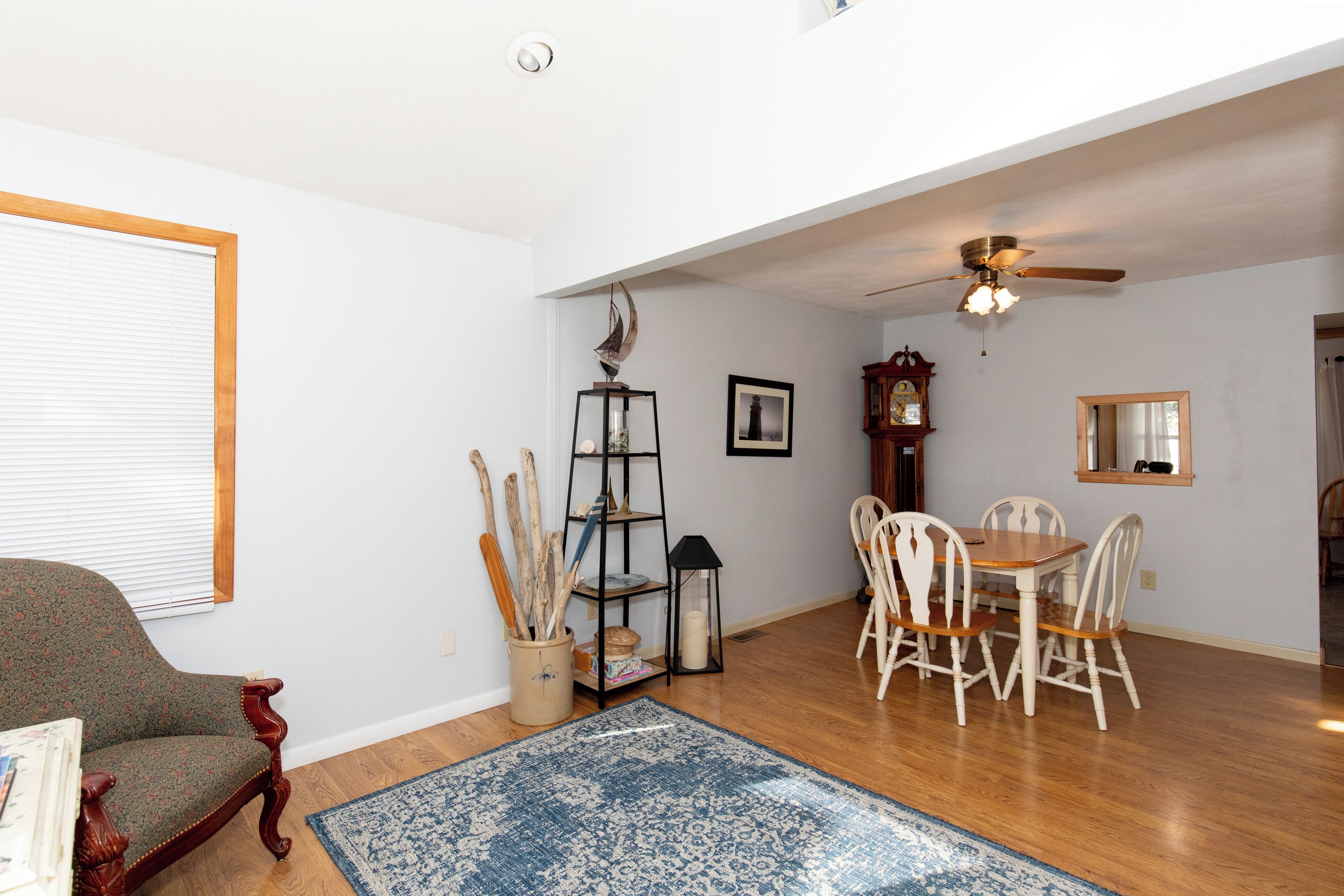 Un salon avec un plancher en bois, une table à manger avec des chaises, un fauteuil en bois, un tapis bleu à motifs, un ventilateur de plafond et une cheminée.