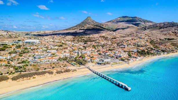An aerial view of a coastal town with a prominent beach, a pier extending into the sea, and a mountain in the background. - Sandybeachapartment1 in the Golden Island (Vila Baleira)