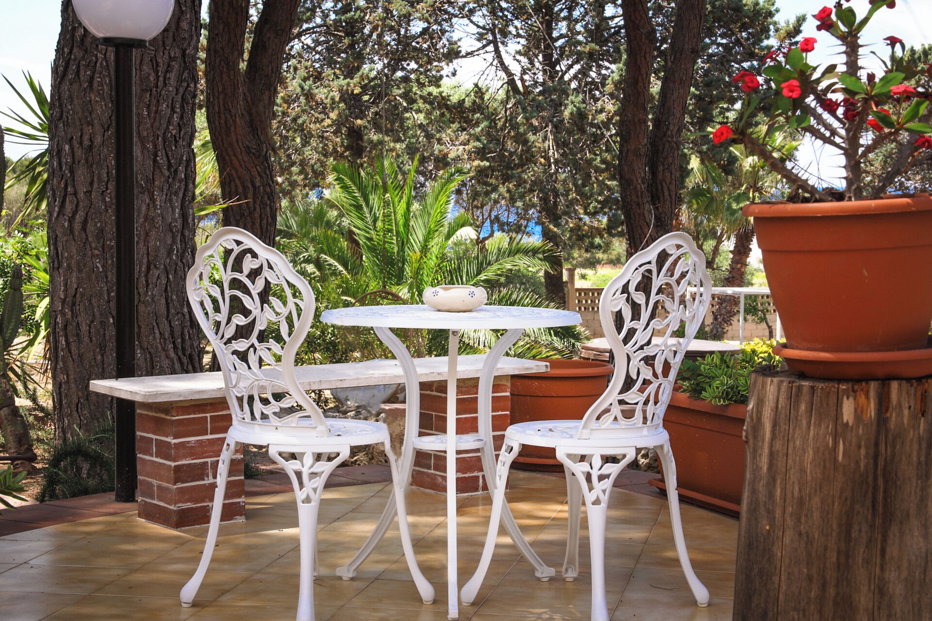 An outdoor patio with white metal chairs and a round table, surrounded by trees and a large terracotta pot.