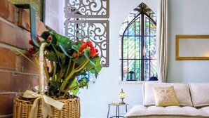 Ett rum med en korg av rotting som innehåller röda blommor, en bekväm soffa med en dekorativ kudde, ett litet bord med en lampa och ett fönster med målat glas.