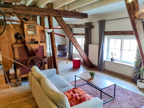 Ein rustikaler Raum mit freiliegenden Holzbalken, einer alten Maschine, einem Sofa, einem roten Hocker und einem Fenster mit Vorhängen.