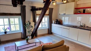 Una cocina moderna con armarios blancos, encimera negra y pileta de acero inoxidable. Se observa una escalera de madera que conduce a un nivel superior, y un sofá color beige en la sala.