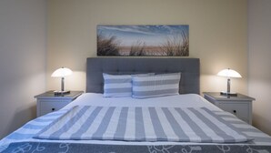 A neatly made bed with striped bedding, two bedside lamps, and a painting of a beach scene on the wall. - Ferienwohnung in St. Peter-Ording (St Peter-Ording)