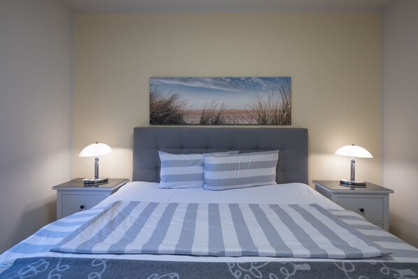 A neatly made bed with striped bedding, two bedside lamps, and a painting of a beach scene on the wall.