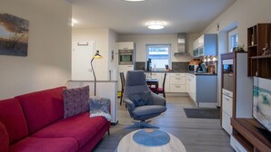 A modern living room with a red sofa, a grey armchair, and a round coffee table. A kitchen with white cabinets and a window is visible in the background. - Ferienwohnung in St. Peter-Ording (St Peter-Ording)