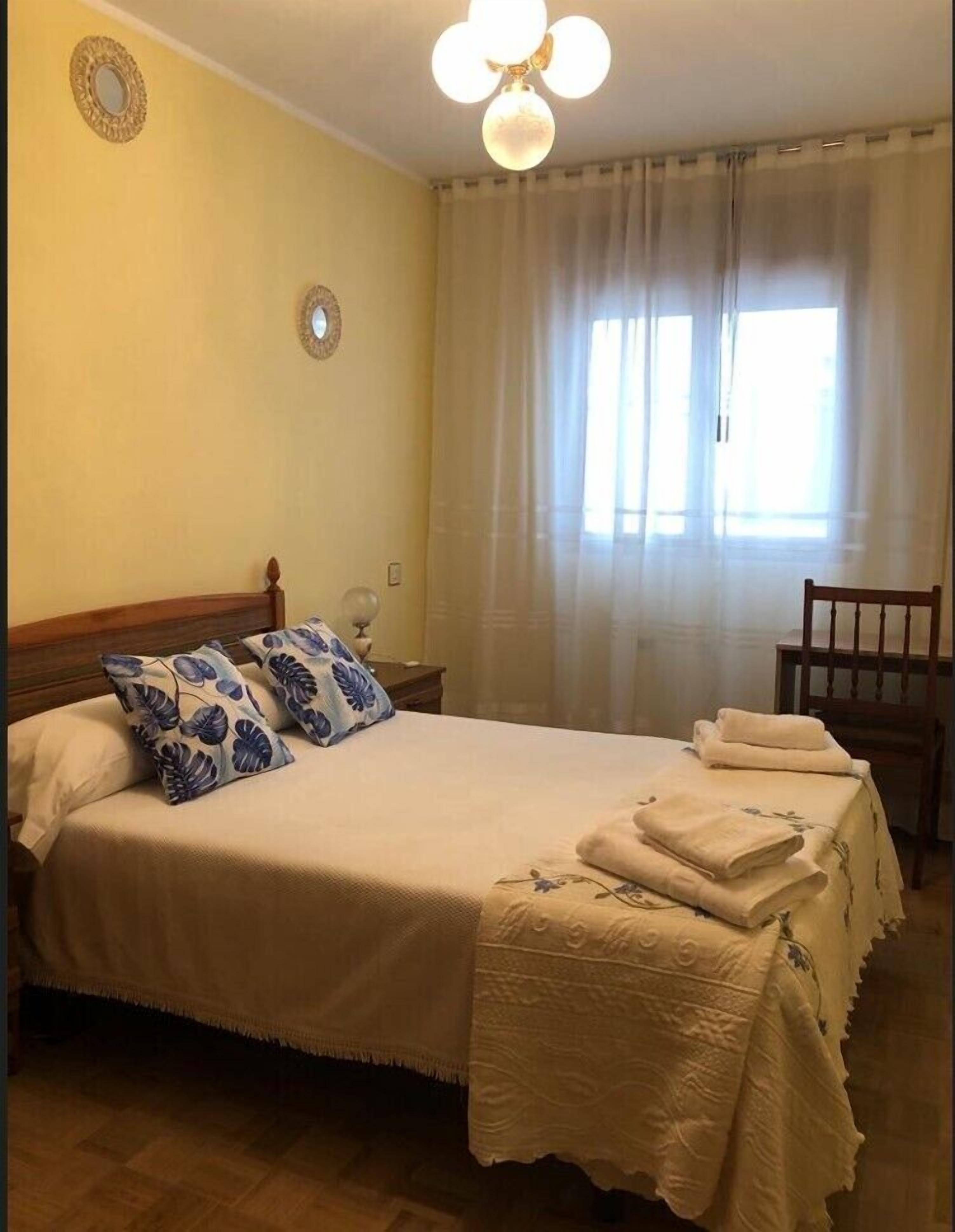 Ein ordentlich bezogenes Bett mit blumigen Kissen, einer Nachttischlampe und einem Sessel.