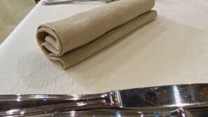 Una mesa de comedor con un mantelino beige doblado, cubiertos y copas de vino, en una habitación con paredes de piedra y techo de madera.