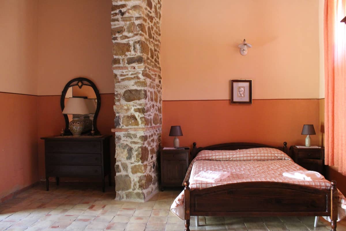 A bedroom with a stone wall, a wooden bed, two bedside tables with lamps, and a dresser.