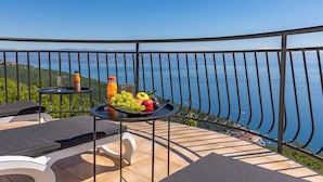 Un balcon avec une table dressée, ornée de fruits et d’une bouteille, offrant une vue panoramique sur la mer et les collines.