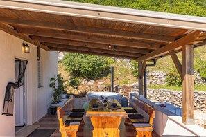 Un espacio comedor al aire libre techado, con una mesa y bancos de madera, un muro de piedra y una puerta con mosquitera.