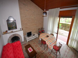 A living room with a fireplace, a television, a dining table with chairs, and a view through a glass door. - Villa With Garden in Lignano Riviera (Lignano Sabbiadoro)