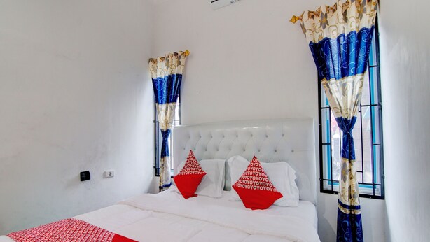 A bedroom with a white bed, red and white patterned pillows, and blue and white curtains. - Hotel O Raya Guest House Syariah (Pekanbaru)