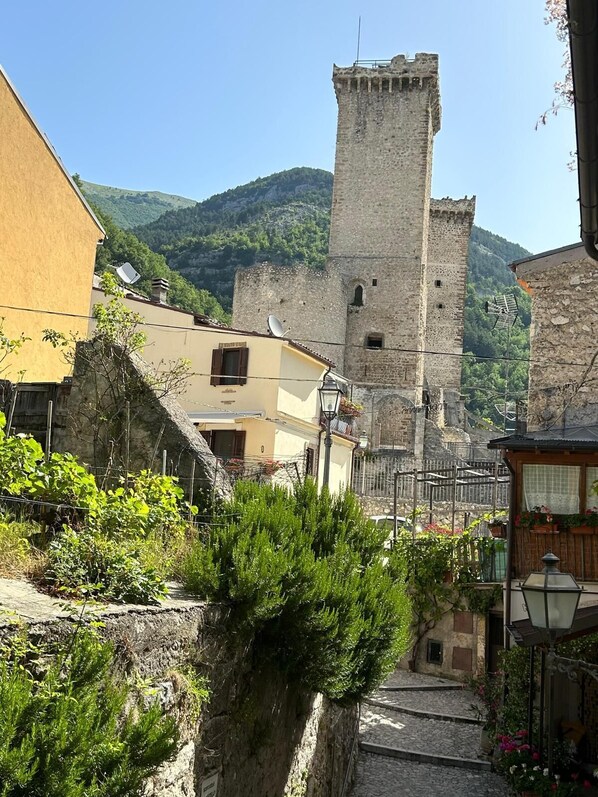 A stone tower with a narrow staircase leading up to it, surrounded by buildings with balconies and potted plants.