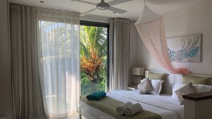 A hotel room with a bed, curtains, a ceiling fan, a nightstand, a lamp, and a view of palm trees. - Appartement De Charme, West Island, Rivière Noire (Black River)