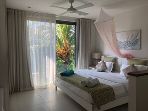 Habitación de hotel con cama, cortinas, ventilador de techo, mesita de noche, lámpara y vista a palmeras.