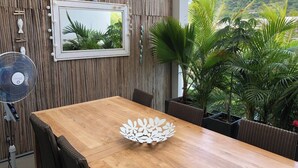 A dining area with a wooden table, wicker chairs, a wall-mounted mirror, and a fan. - Appartement De Charme, West Island, Rivière Noire (Black River)