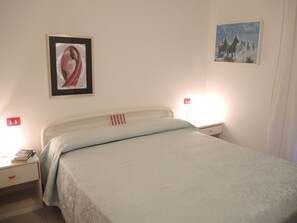 A bedroom with a bed, two bedside tables, a nightstand, a framed picture on the wall, and a lamp on the nightstand. - Pool and Beach Apartment in Bibione Spiaggia (San Michele al Tagliamento)