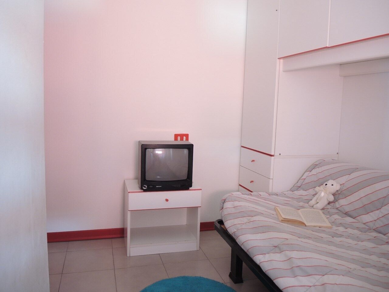 A bedroom with a bed, a nightstand with a television, and a teddy bear on the bed.