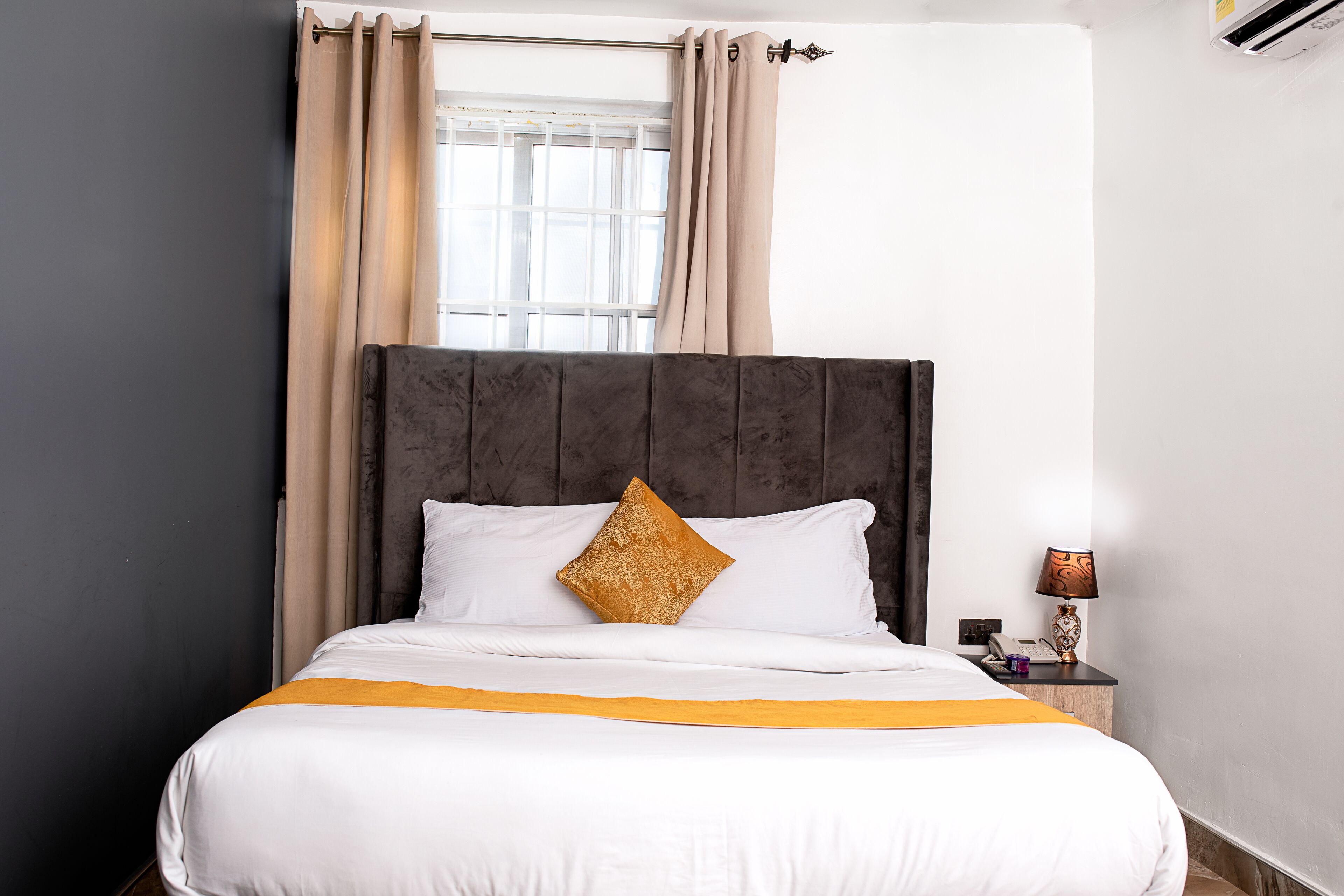 A neatly made bed with white linens and a gold accent pillow, a bedside table with a lamp, and a window with curtains.