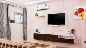Una sala de estar moderna con un televisor de pantalla plana grande montado en un mueble de madera, un sofá capitonado, una cortina estampada y un ventanal con vistas al exterior.