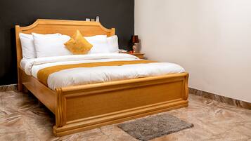A wooden bed with white bedding and a mustard-colored throw, a nightstand with a lamp, and a marble floor.