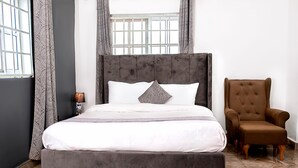 Una cama con cabecera gris tapizada, ropa de cama blanca y una sola almohada gris. Un sillón marrón está situado a la derecha de la cama.