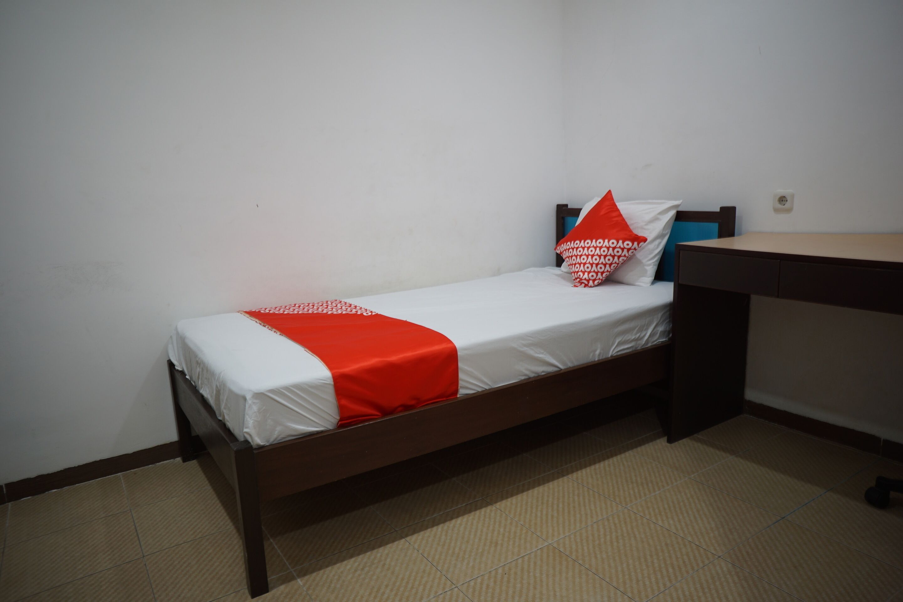 A single bed with a white and red bedspread, a wooden headboard, and a small desk with a chair.